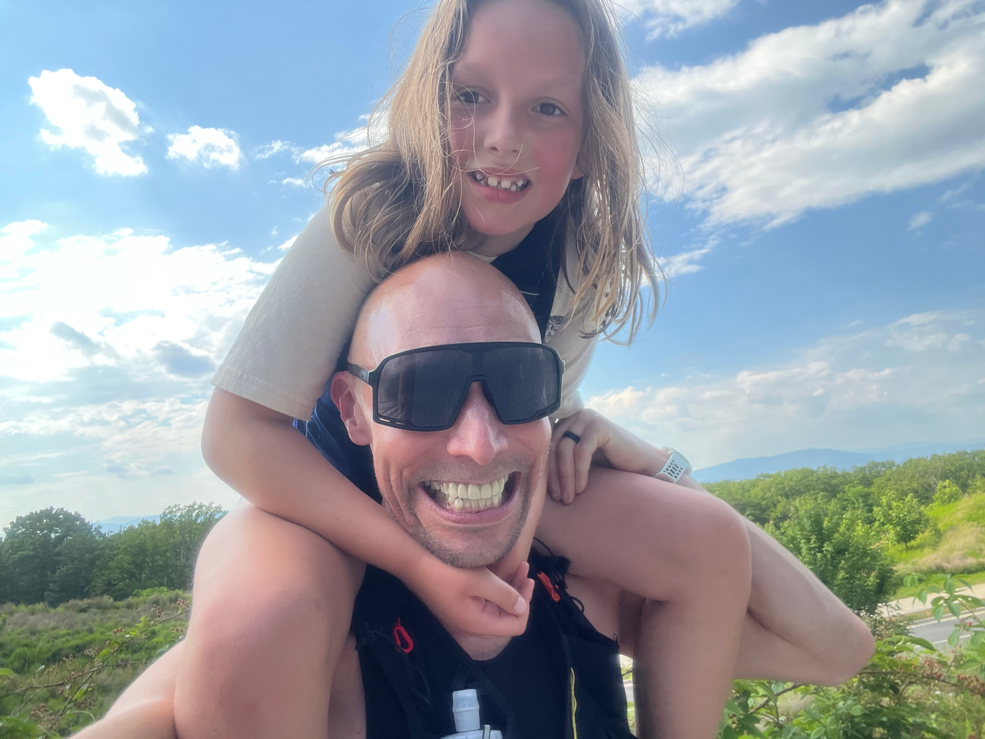 Tim and Amelia Scott outdoors, with Amelia riding on Tim's shoulders against a bright blue sky.